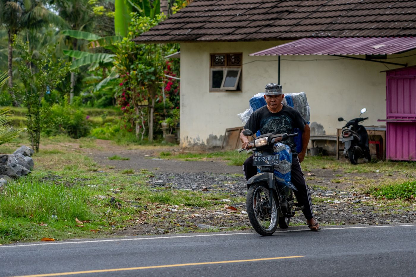 A rider carrying goods on a motorcycle past a village house